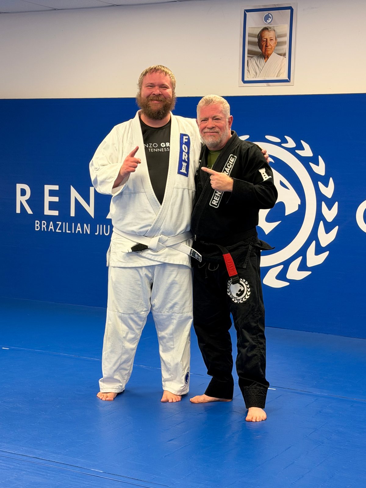 Two men in white and black Brazilian jiu-jitsu uniforms pose on blue mats beneath a framed portrait.