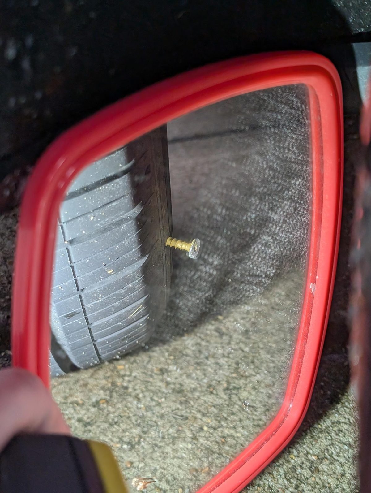 Close-up of a red-framed vehicle mirror reflecting a tire, with a screw protruding near the tire.