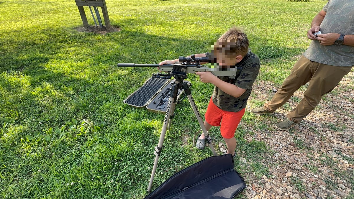 Boy aiming a scoped rifle on a tripod while an adult stands beside him on a grassy field.