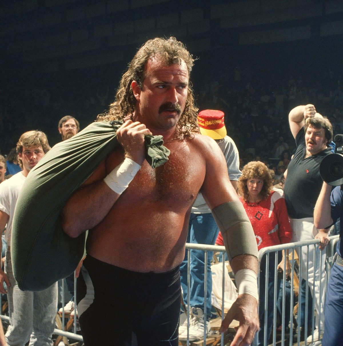Wrestler with a bag over his shoulder in an arena, with spectators behind a barrier.