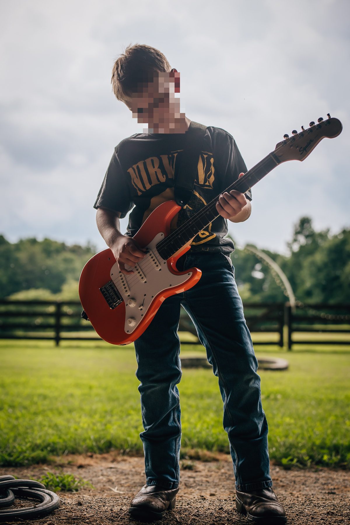 Boy in jeans and boots plays an orange electric guitar outdoors, wearing a Nirvana shirt.