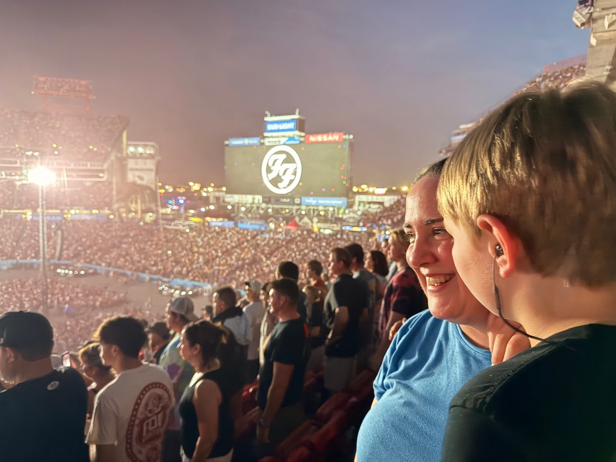 Crowd gathered at night beneath a lit sign, with a smiling woman in a blue shirt in the foreground.