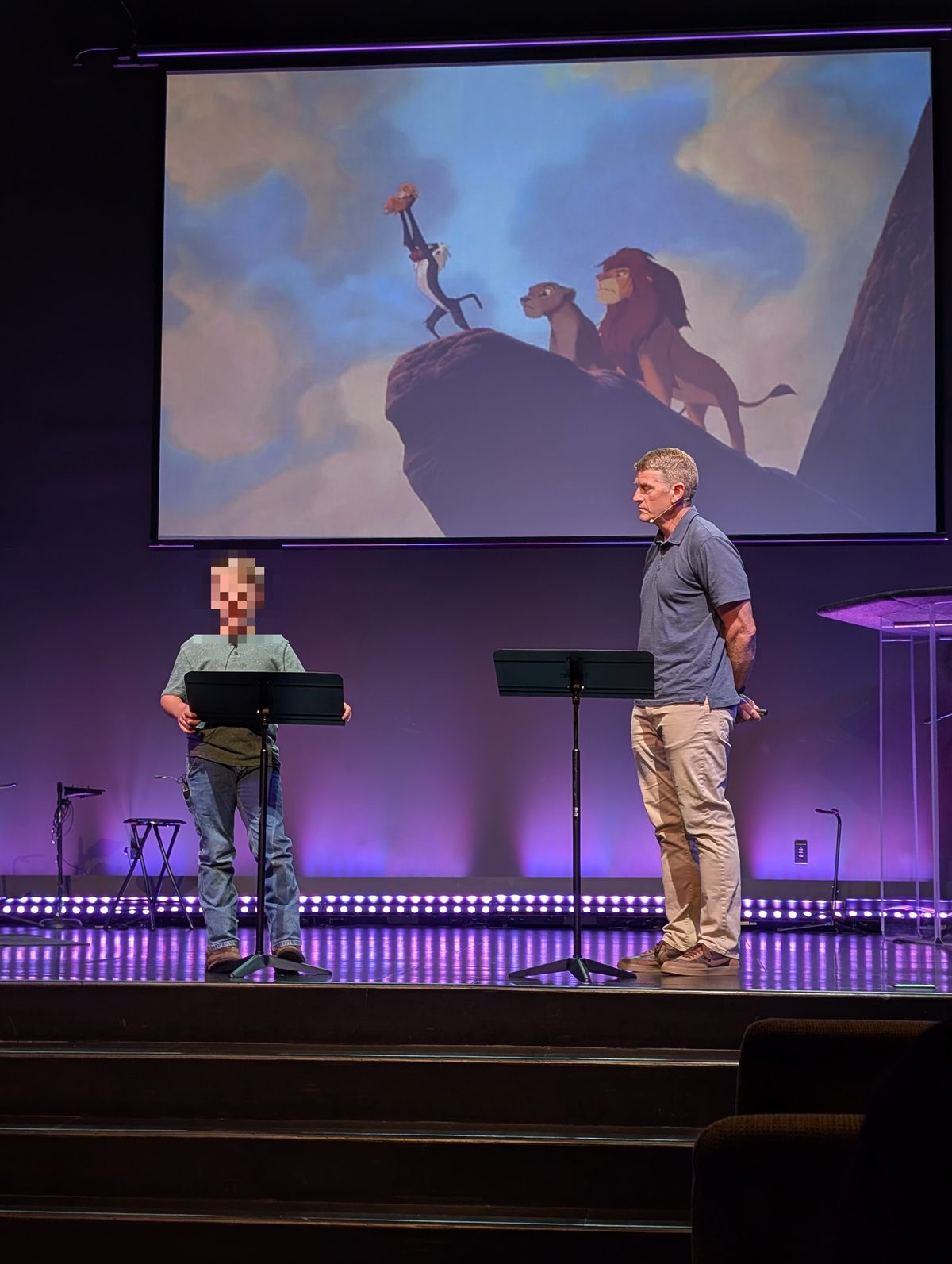 Boy and man on a stage behind lecterns, with a projected Lion King scene above them.