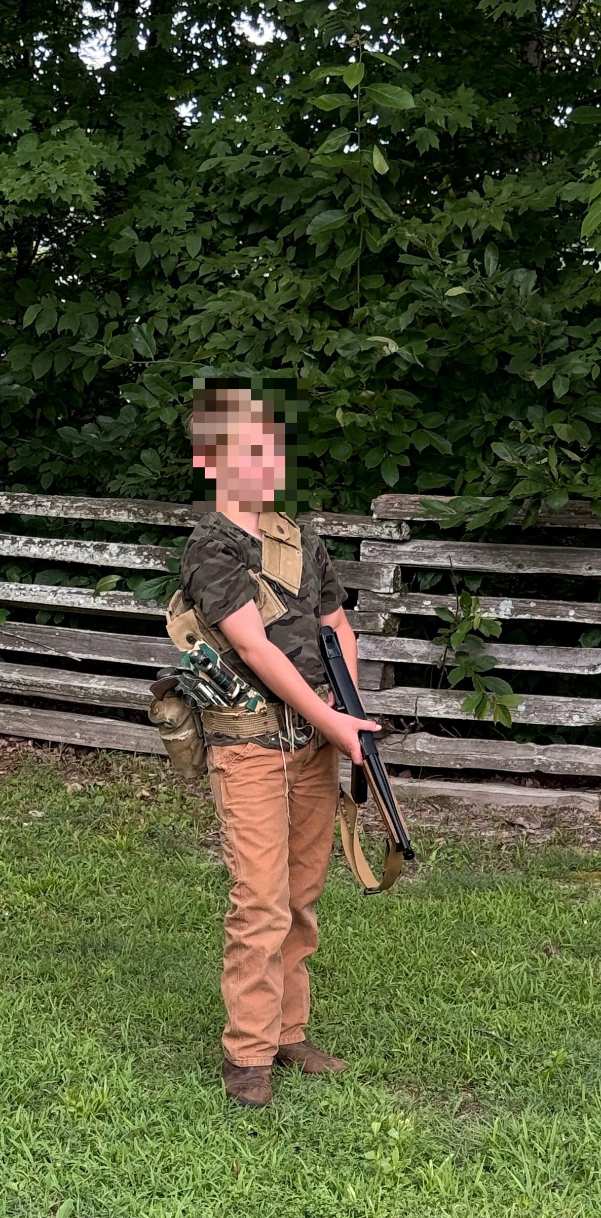 Boy in camouflage holds a rifle outdoors beside a weathered wooden fence and dense trees.