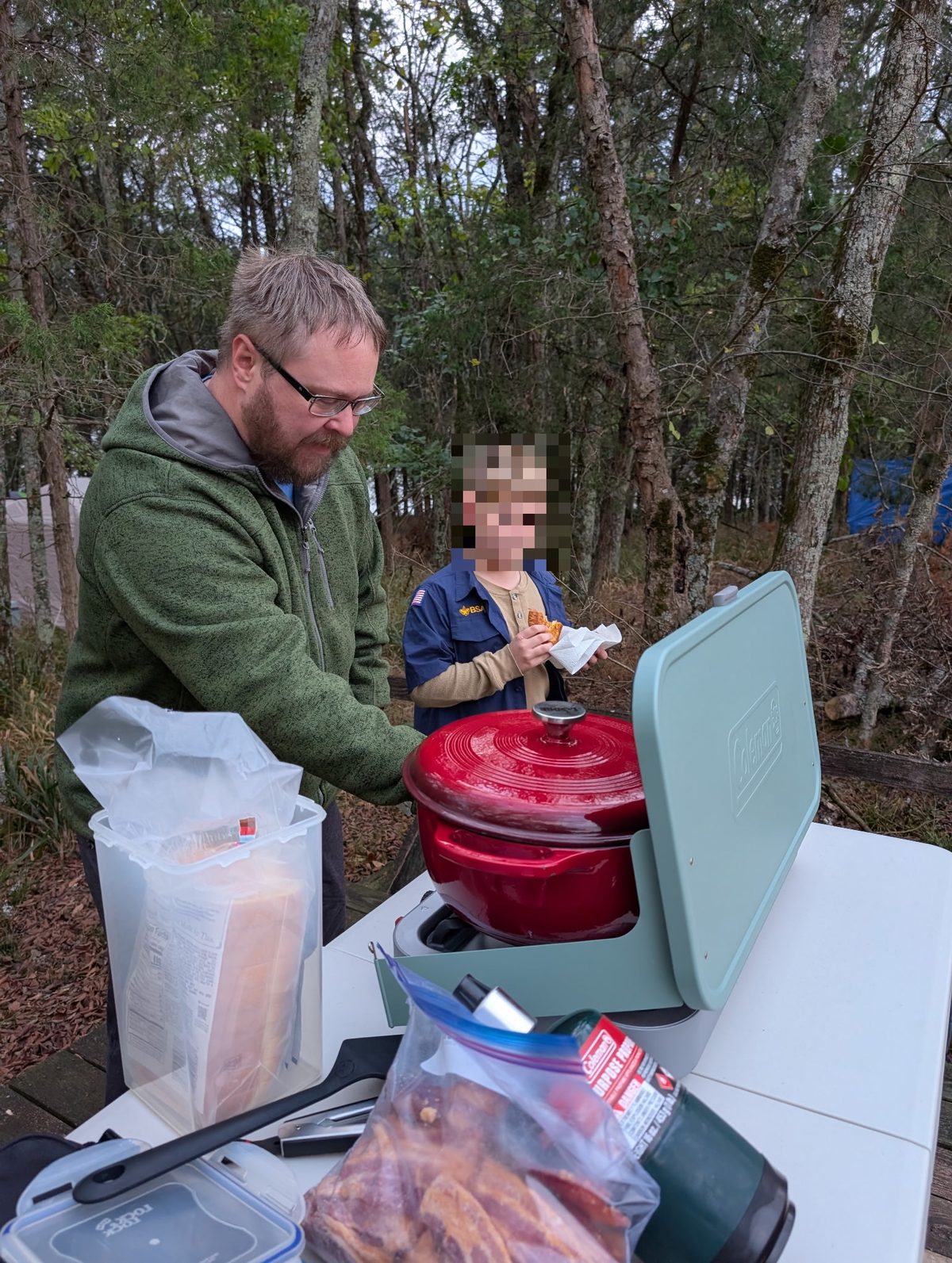 Man and child beside a red lidded grill, with food and cooking supplies on a table in a wooded area.