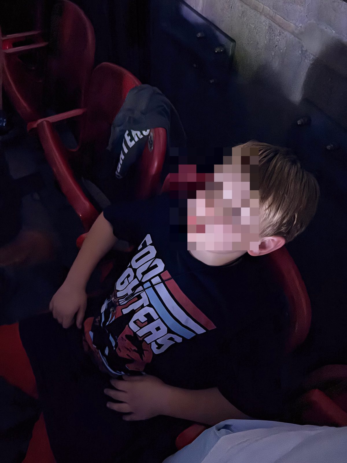 Child seated in a dark theater seat, wearing a black graphic shirt, with red seats and a wall behind.