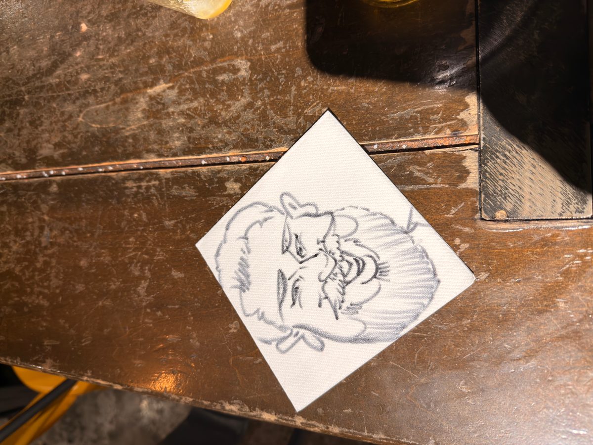 Pencil sketch of a bearded man’s face on a napkin on a worn wooden table.
