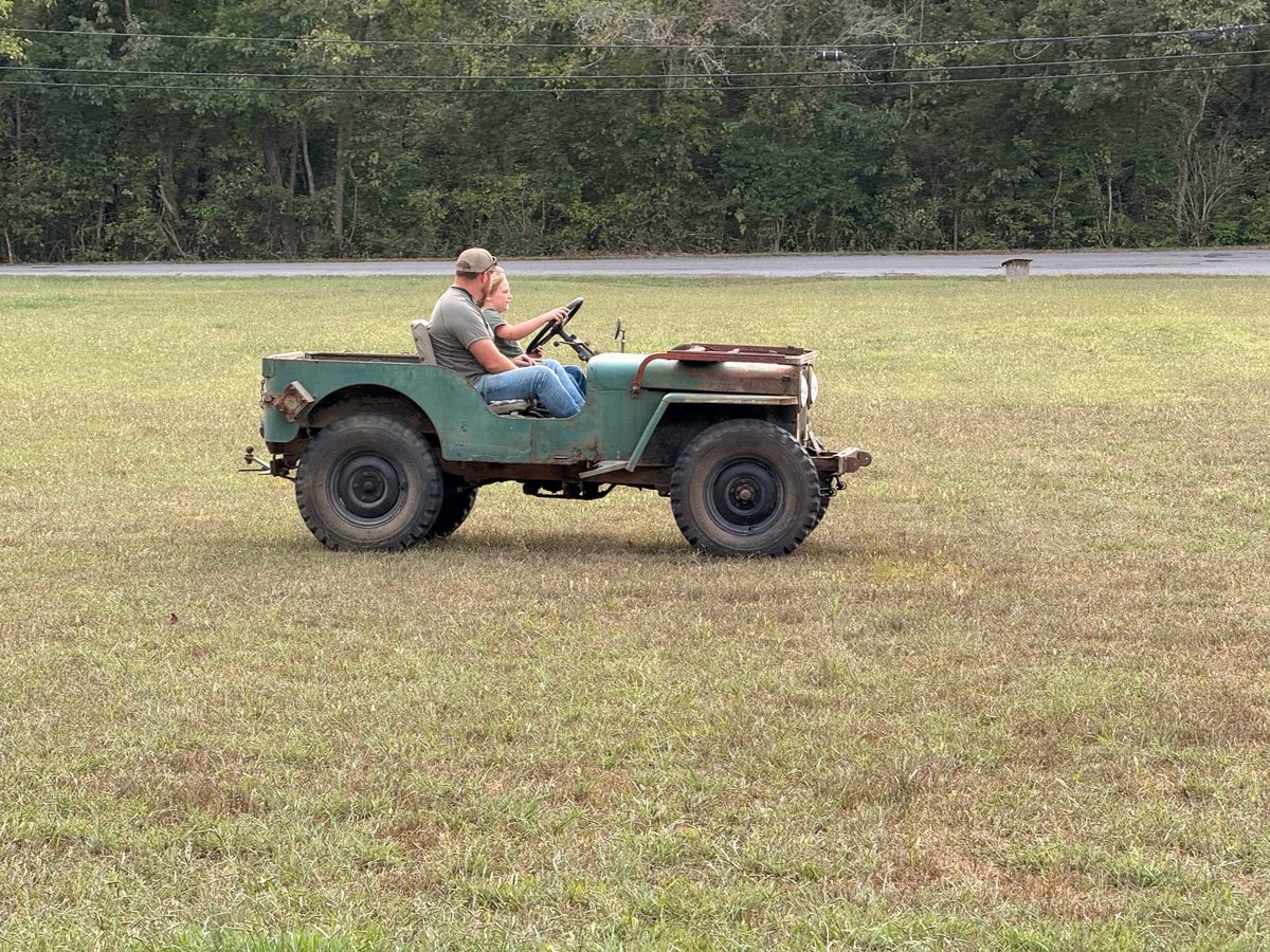 Two people ride in a weathered green jeep across a grassy field, with trees, a road, and overhead wires behind them.
