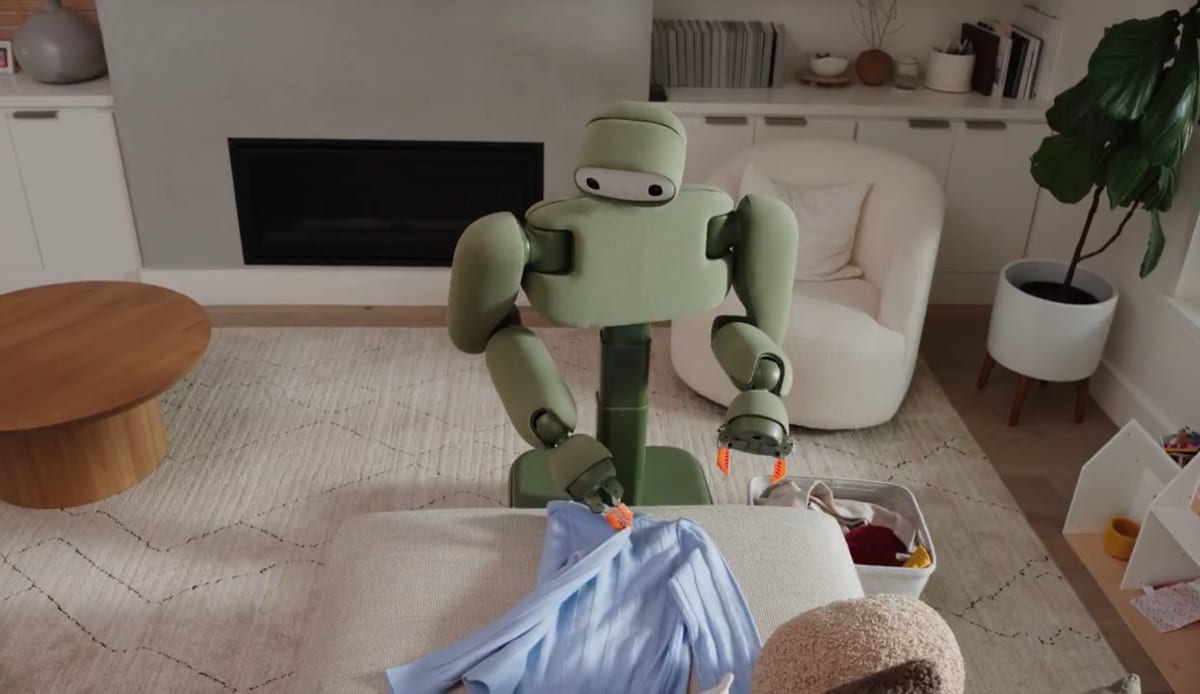 Green humanoid robot stands in a living room folding clothes on a couch ottoman.